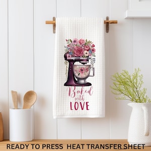 May include: White kitchen towel with a floral design featuring a stand mixer and the words "Baked with Love" in burgundy. The towel is hanging on a wooden rack. A vase with greenery is on the right.