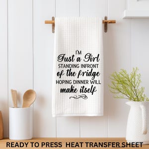 May include: A white kitchen towel with the text "I'M Just a Girl standing in front of the fridge hoping dinner will make itself." The towel is hanging on a wooden rod. A ceramic jar, a white container with wooden utensils, and a white pitcher with greenery are also visible.