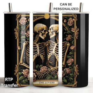 May include: A black tumbler featuring a tarot card design with two skeletons embracing, framed by roses and ornate gold detailing. The text "THE LOVERS" is visible. The tumbler has a silver straw and the text "CAN BE PERSONALIZED" is printed on the tumbler.