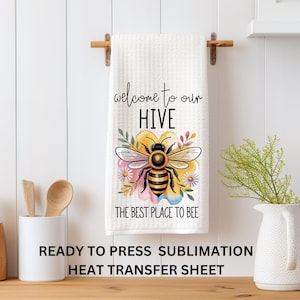May include: White kitchen towel with a bee design and the text "welcome to our HIVE THE BEST PLACE TO BEE". The towel is hanging on a wooden rod. A decorative bee is in the center of the towel, surrounded by flowers.