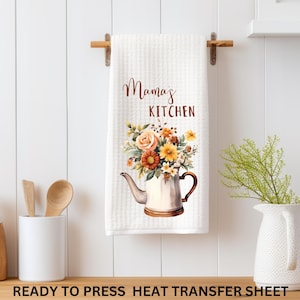 May include: White kitchen towel with the text "Mama's Kitchen" and a floral design in a teapot. The towel is hanging on a wooden rod. A wooden spoon and spatula are in a white container.