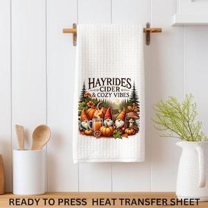 May include: White waffle-weave kitchen towel with a fall-themed graphic. The graphic features the text "HAYRIDES CIDER & COZY VIBES" above an illustration of pumpkins, gnomes, and a mug of cider. The towel is hanging on a wooden rod.