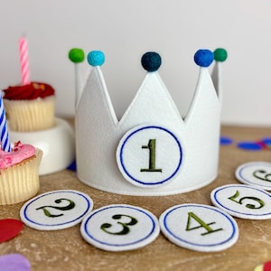 Può includere: Corona in feltro bianco con palline in feltro blu e un numero uno in feltro verde sul davanti. La corona è circondata da cerchi in feltro bianco con bordi in feltro blu e numeri in feltro verde da 2 a 6.