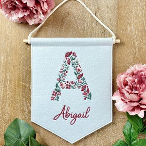 May include: A light blue felt banner with a white rope hanger. The banner features a floral letter A embroidered in shades of pink and green. The name Abigail is embroidered below the letter in pink.