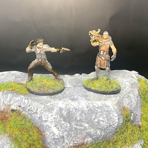 May include: Two painted miniature figures, one holding a gun and the other holding a dragon head, stand on a gray rock with green grass.