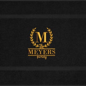 May include: Black rectangular doormat with a gold family crest design. The crest features a gold letter "M" within a laurel wreath, with "the MEYERS family" written below in a script font. The doormat has a textured surface and a dark border.