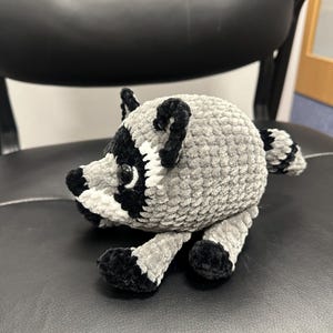 May include: A small, round, crocheted raccoon plush toy made of soft, textured grey yarn. It features a black and white face mask, black ears, black paws, and a striped tail. The raccoon has shiny black plastic eyes and a rounded body shape.
