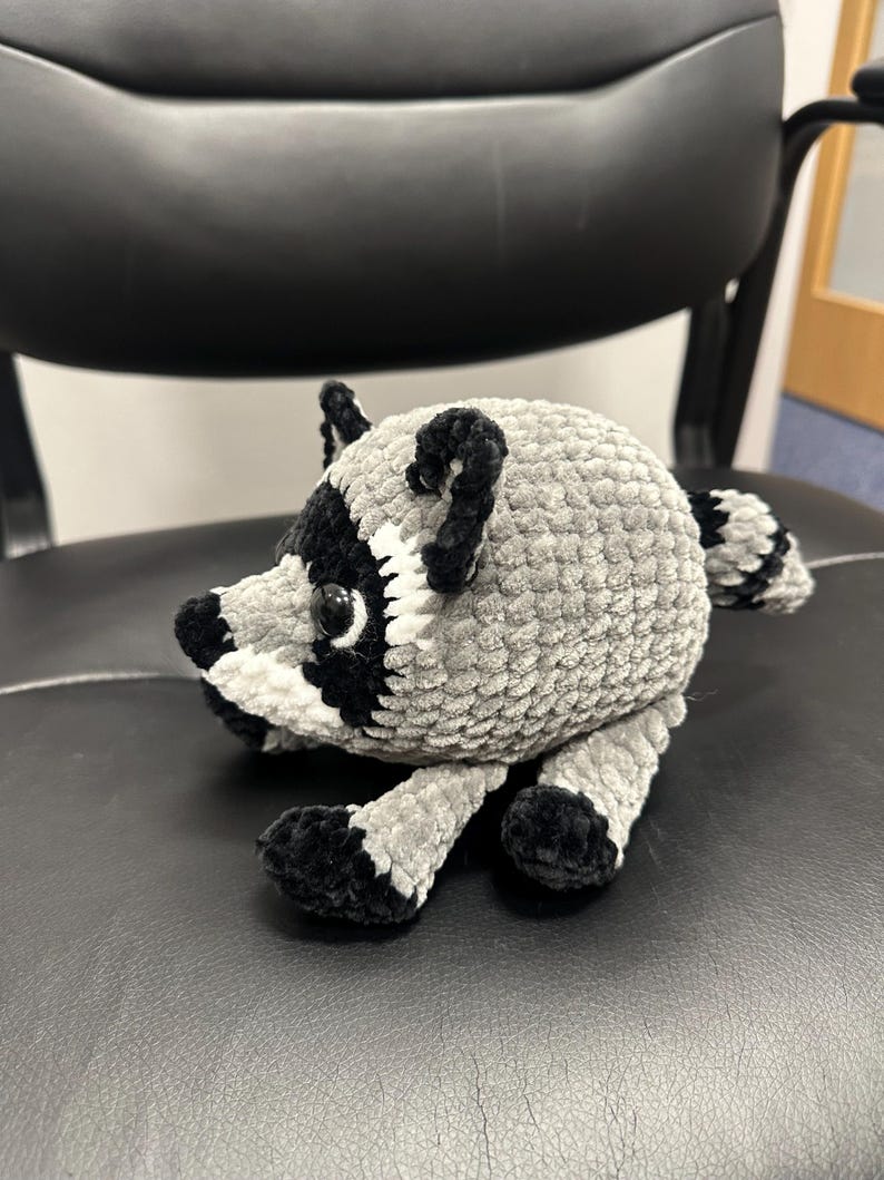 May include: A small, round, crocheted raccoon plush toy made of soft, textured grey yarn. It features a black and white face mask, black ears, black paws, and a striped tail. The raccoon has shiny black plastic eyes and a rounded body shape.