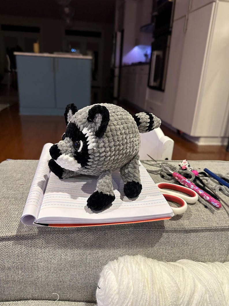 May include: A round, plush, crocheted raccoon toy made of soft grey yarn with black and white facial markings, black ears, and a striped tail. The stuffed animal stands on a white lined notebook next to a pair of scissors and a crochet hook.