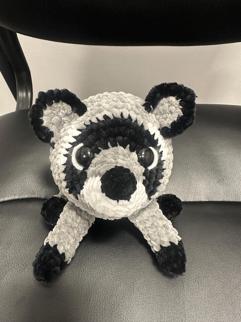 May include: A plush raccoon toy made of soft, fuzzy yarn in shades of gray, black, and white. The stuffed animal features large, round, glossy black eyes, a black nose, and distinct black and white facial markings with rounded ears.