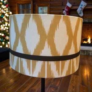 Può includere: Paralume cilindrico con sfondo color crema e motivo a rombi dorati ripetuti. Una fascia marrone scuro circonda il paralume. La lampada è su un supporto nero, con un albero di Natale e un camino sullo sfondo.