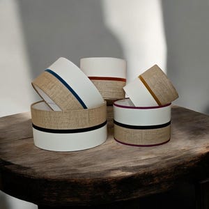 Può includere: Una collezione di paralumi cilindrici, con sezioni bianche, fasce in juta e strisce colorate. I paralumi sono disposti su una superficie di legno rustico. Il design combina elementi moderni con texture naturali.