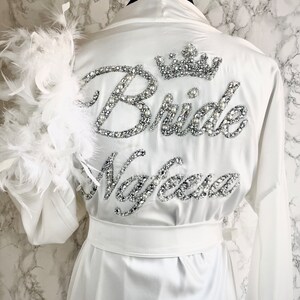 May include: White satin robe with a feather trim on the sleeves. The robe has a rhinestone embellishment on the back that says 'Bride' with a crown above it and 'Maylee' below it.