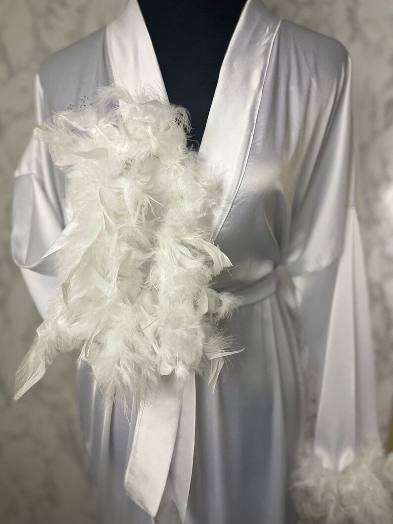 May include: A white satin robe with a white feather trim around the collar and cuffs. The robe has a sash tie at the waist and a large white feather accent on the left side of the chest.