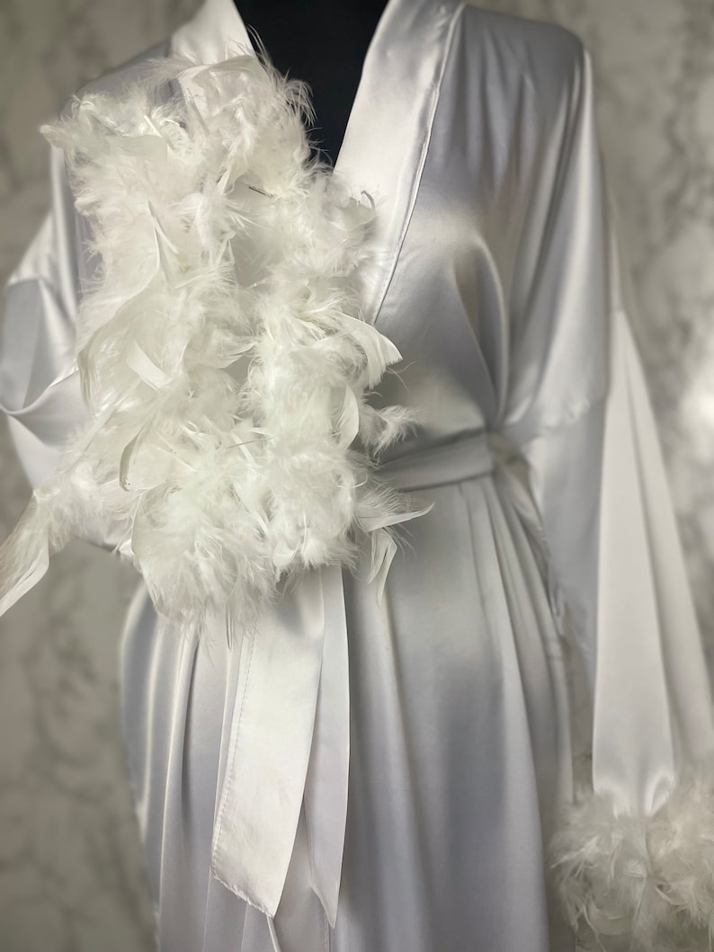 May include: A white satin robe with a white feather trim around the neckline and cuffs. The robe has a sash tie at the waist.