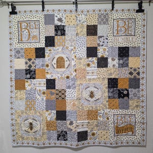 May include: A square patchwork quilt featuring a color palette of yellow, black, white, and gray. The quilt includes square panels with bee, beehive, and floral motifs, along with text panels that read Bee Kind, Be Humble, and Bee Joyful.