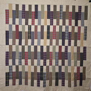 May include: A rectangular quilt featuring a patchwork design of vertical fabric strips in various plaid and checkered patterns. The colors include shades of red, blue, green, brown, and tan, all set against a light beige background fabric.