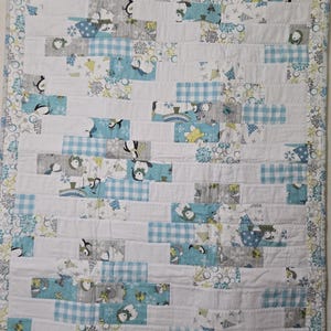 May include: A rectangular quilt featuring a patchwork design of horizontal fabric strips in shades of white, light blue, and gray. The fabric patterns include illustrations of penguins, snowflakes, and blue gingham checks, finished with a patterned border.
