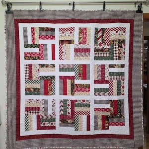 May include: A quilt featuring a rail fence block pattern with various holiday-themed fabrics in red, green, and white. The quilt has a wide red inner border and a dark brown and white checkered outer border, all finished with white stitching.