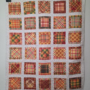 May include: A rectangular quilt featuring a grid of thirty square fabric patches in various plaid patterns of orange, yellow, red, green, and brown. The squares are separated by a cream-colored fabric with a subtle, swirling quilted texture.