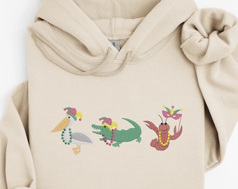 May include: Beige hoodie with embroidered Mardi Gras-themed designs. The designs include a pelican, an alligator, and a crawfish, each wearing a festive hat and beads. The crawfish holds a mask. The hoodie is made of a soft material.