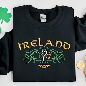 K&ouml;nnte beinhalten: Schwarzer Pullover mit einem goldenen gestickten Kleeblatt und dem Wort "IRELAND" in goldenen Buchstaben.
