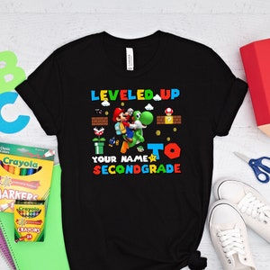 May include: Black t-shirt with a video game character graphic. The text "LEVELED UP TO SECONDGRADE" is printed on the shirt.