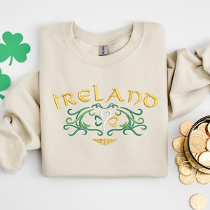 K&ouml;nnte beinhalten: Ein beiger Pullover mit einem goldenen Stickmuster, auf dem "IRELAND" mit einem Kleeblatt in der Mitte steht. Das Kleeblatt ist von gr&uuml;nen Ranken umgeben.