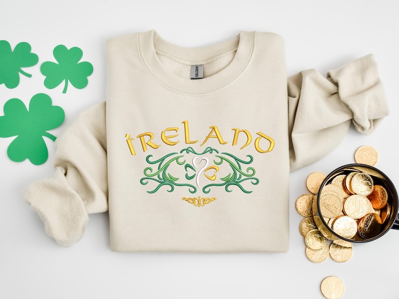 K&ouml;nnte beinhalten: Ein beiger Pullover mit einem goldenen Stickmuster, auf dem "IRELAND" mit einem Kleeblatt in der Mitte steht. Das Kleeblatt ist von gr&uuml;nen Ranken umgeben.