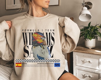 May include: Beige Formula 1 Team sweatshirt featuring a graphic of a driver in a helmet. The text reads "WILLIAMS SAINZ" in black block letters. Below the text is a checkered flag design with the text "SMOOTH OPERATOR".