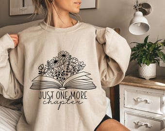 May include: Beige sweatshirt with a black graphic of an open book with flowers and the text "JUST ONE MORE chapter". The design features daisies and other floral elements. A cozy, casual top for book lovers.