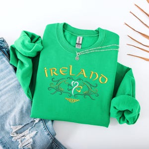 K&ouml;nnte beinhalten: Gr&uuml;ner Pullover mit dem Wort "IRELAND" in goldenen Buchstaben und einem stilisierten Kleeblatt-Design. Der Pullover liegt auf einer wei&szlig;en Oberfl&auml;che mit einer blauen Jeans.