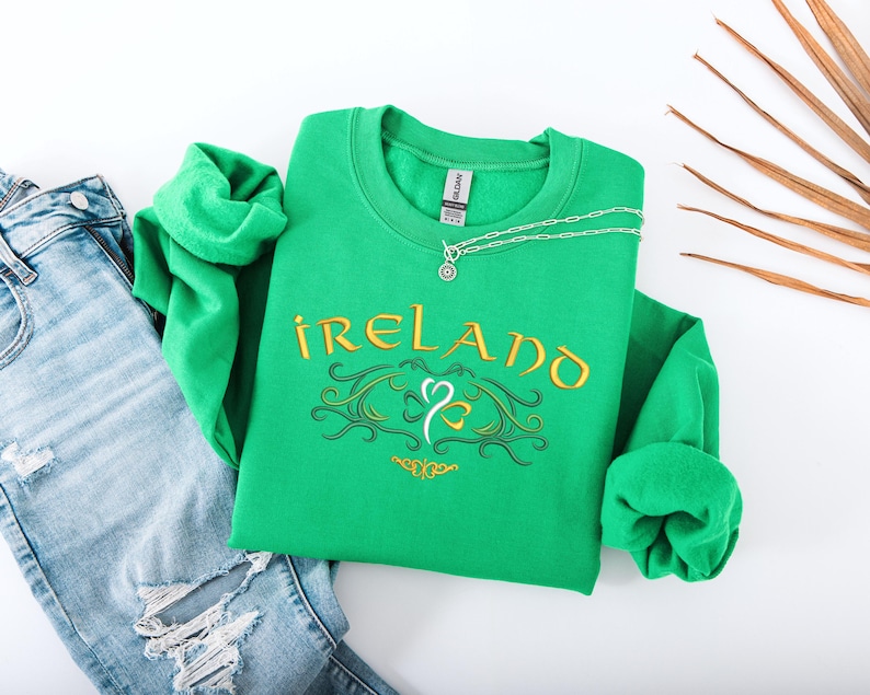 K&ouml;nnte beinhalten: Gr&uuml;ner Pullover mit dem Wort "IRELAND" in goldenen Buchstaben und einem stilisierten Kleeblatt-Design. Der Pullover liegt auf einer wei&szlig;en Oberfl&auml;che mit einer blauen Jeans.
