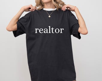 May include: Black oversized t-shirt with the word "realtor" printed in white. The shirt has a white collar and is being held up by a person. The shirt is made of a soft material.
