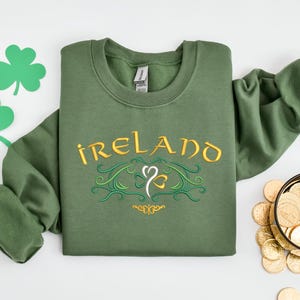 K&ouml;nnte beinhalten: Gr&uuml;nes Sweatshirt mit dem Wort "IRELAND" in goldener Schrift und einem Kleeblatt-Design bestickt.