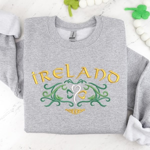 K&ouml;nnte beinhalten: Ein grauer Pullover mit dem Wort "IRELAND" in goldenen Buchstaben und einem gr&uuml;nen Kleeblatt-Design gestickt.