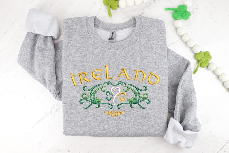 K&ouml;nnte beinhalten: Ein grauer Pullover mit dem Wort "IRELAND" in goldenen Buchstaben und einem gr&uuml;nen Kleeblatt-Design gestickt.