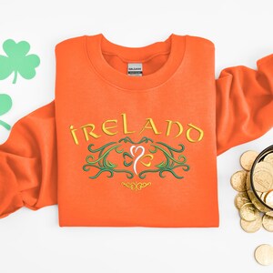 K&ouml;nnte beinhalten: Orangefarbener Pullover mit einem gestickten Design in Gr&uuml;n und Gold, auf dem "IRELAND" mit einem Kleeblatt in der Mitte steht.
