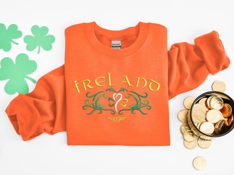 K&ouml;nnte beinhalten: Orangefarbener Pullover mit einem gestickten Design in Gr&uuml;n und Gold, auf dem "IRELAND" mit einem Kleeblatt in der Mitte steht.