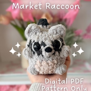 May include: A crocheted raccoon toy in shades of gray and beige, with large black eyes and a black nose. The text "Willow Forest Market Raccoon" and "Digital PDF Pattern Only" are displayed. The creator's name, "kybeecreates", is also visible.