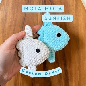 May include: Two crocheted sunfish, one white and one light blue, with black button eyes. The text "MOLA MOLA SUNFISH" and "Custom Order" are displayed on the image. The sunfish are held against a wooden background.