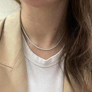 May include: A delicate, sparkling necklace with numerous small, clear stones. The necklace is worn against a white ribbed shirt and a tan blazer. The stones appear to be evenly sized and closely set, creating a continuous line of brilliance.