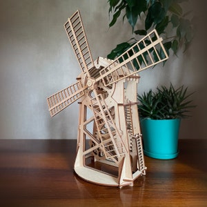 May include: A wooden model of a windmill with intricate details and working gears. The model is made of light brown wood and has a white base. The windmill has four blades and is set against a light brown background.