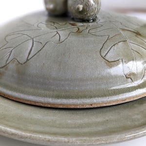 May include: A ceramic butter dish with a lid featuring an oak leaf design and two acorns. The dish is glazed in a light green color.