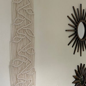 Peut inclure: Une suspension murale en macramé blanche avec un motif géométrique. La suspension est faite de cordon de coton épais et est fixée à un mur blanc. Il y a deux miroirs soleil dorés sur le mur derrière la suspension.