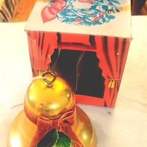 May include: A gold-colored metal bell ornament featuring a brown velvet ribbon bow and green holly leaves. The bell has a metal loop at the top for hanging. It is positioned in front of a cardboard box labeled Musical Christmas Bell.