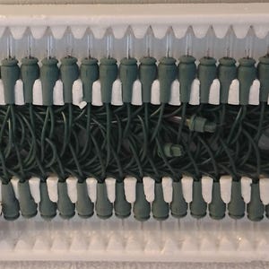 May include: A set of Christmas lights in a white styrofoam tray. The lights have green bases and white bulb holders. The wires are dark green, and the plug is green. A red fuse is attached.