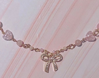 May include: A delicate gold chain necklace with pink and light blue beads, heart charms, and a sparkling bow pendant.