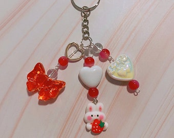 May include: A keychain with a silver ring and chain. It features a red bow, a gold heart, white and iridescent hearts, red beads, clear beads, and a white bunny holding a strawberry. The keychain is a charming accessory.