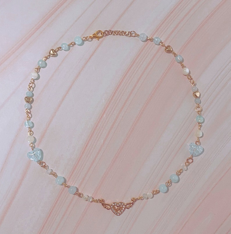 May include: A delicate gold chain necklace with light blue and white beads and a gold heart charm with clear rhinestones.